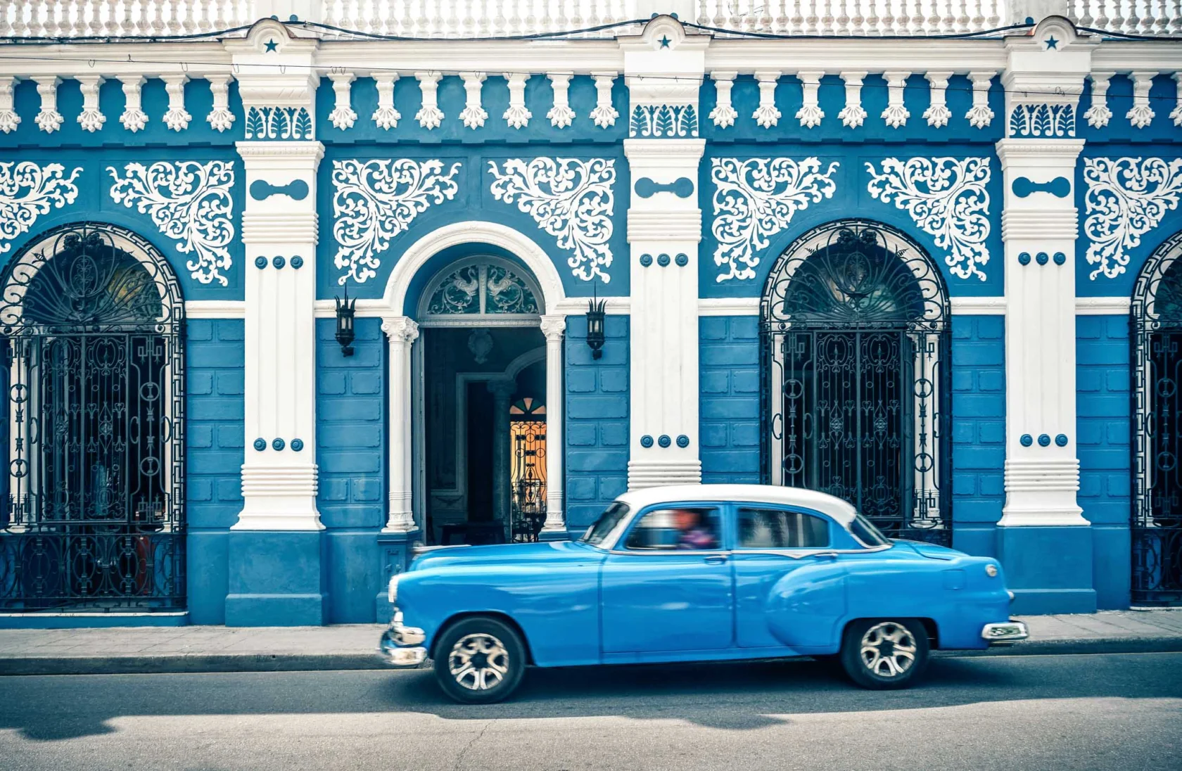 A vintage blue car passes by a blue and white ornate building with arched doorways and decorative wrought iron windows.