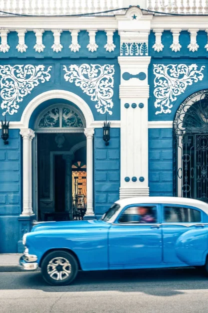 A vintage blue car passes by a blue and white ornate building with arched doorways and decorative wrought iron windows.