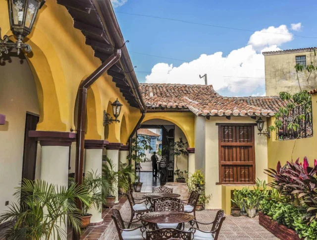 Outdoor courtyard with yellow arches, brown tiled floor, wrought iron furniture, and potted plants, set under a blue sky with clouds.