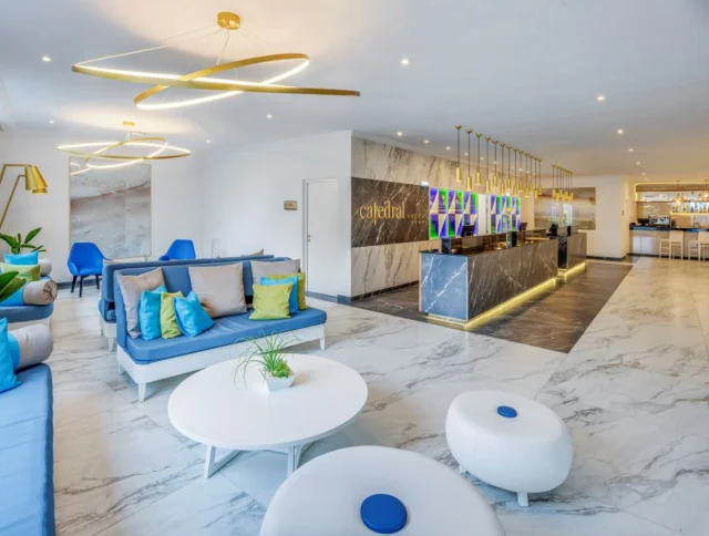 Modern hotel lobby with marble floors, blue and white seating, a sleek black and gold reception desk, and decorative lighting fixtures. A bar is visible in the background.