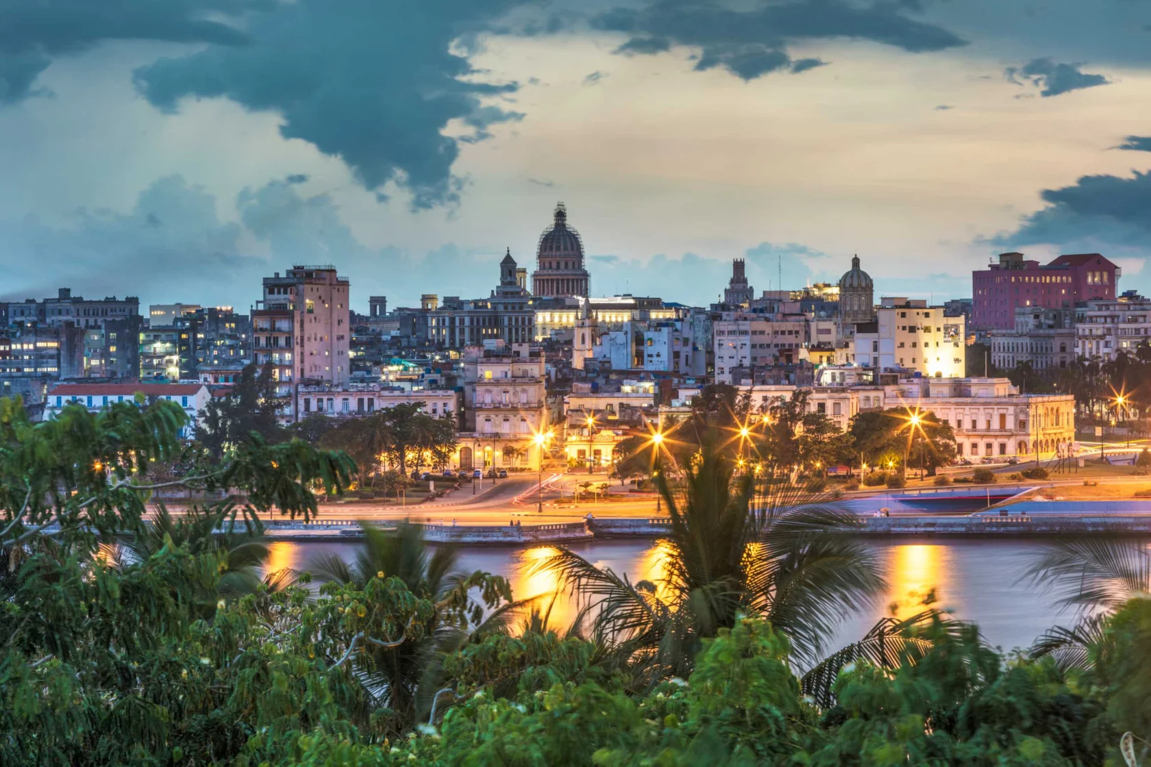 Cityscape of Havana, Cuba at dusk with illuminated buildings, including the Capitolio, and a waterfront reflecting lights in the foreground, framed by lush greenery.