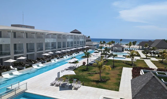 Aerial view of a modern resort with multiple pools, surrounded by loungers and palm trees, set by the ocean under a clear blue sky.