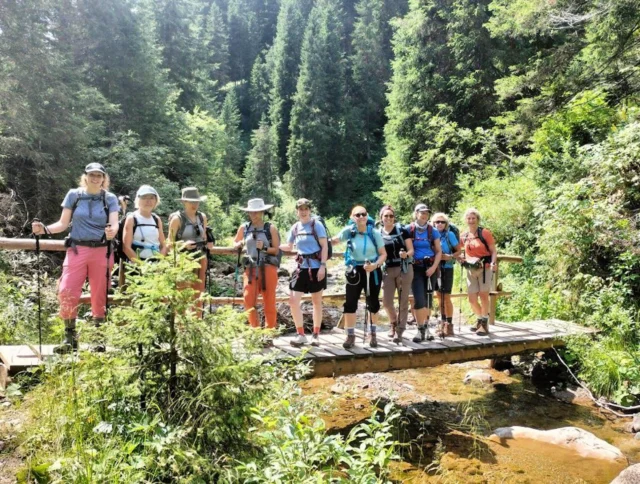 A group of hikers stands on a small wooden bridge in a lush forested area. They are equipped with backpacks and hiking gear.