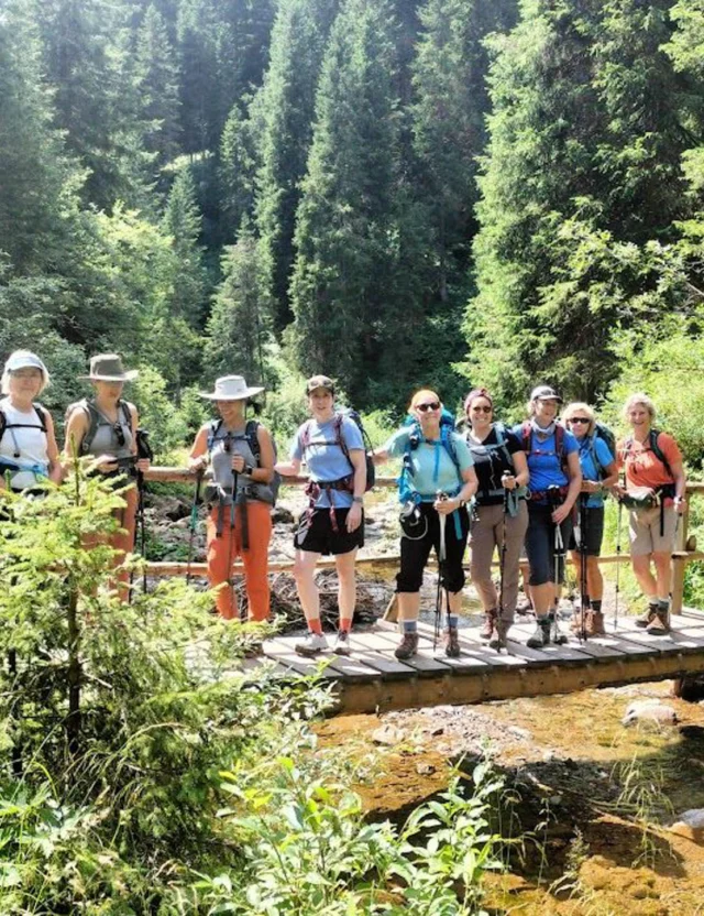 A group of hikers stands on a small wooden bridge in a lush forested area. They are equipped with backpacks and hiking gear.