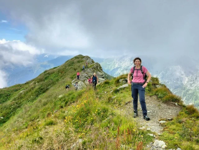 Person in a pink shirt hiking on a narrow trail along a grassy mountain ridge, with several other hikers in the background and a cloudy sky above.