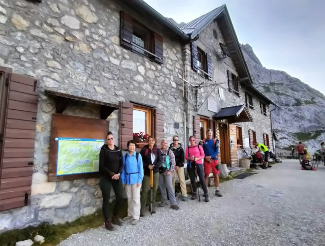 A group of hikers stands outside a stone building on a mountain trail. The sky is slightly overcast, and a map is displayed on the wall.