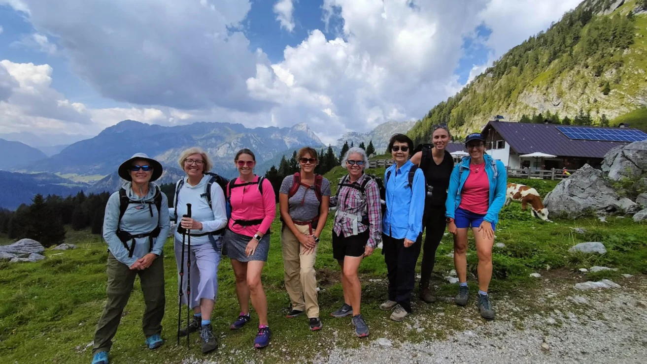 A group of eight women stands outdoors on a hiking trail with mountains and a cabin in the background.