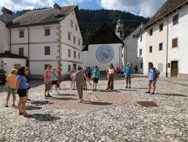 A group of people stand in a circle in a cobblestone plaza surrounded by old buildings and mountains.