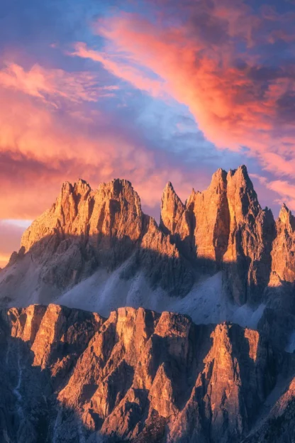 A mountain range with jagged peaks is bathed in orange and pink light from a vibrant sunset sky.