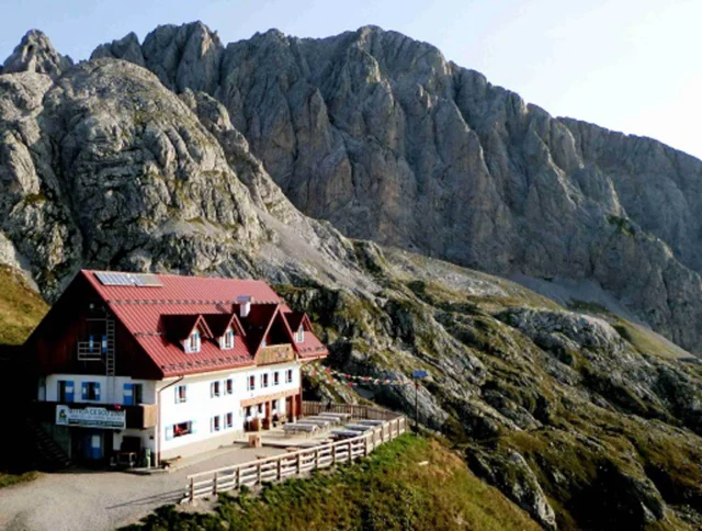 A large mountain lodge with a red roof is situated at the base of rocky cliffs under a clear sky.