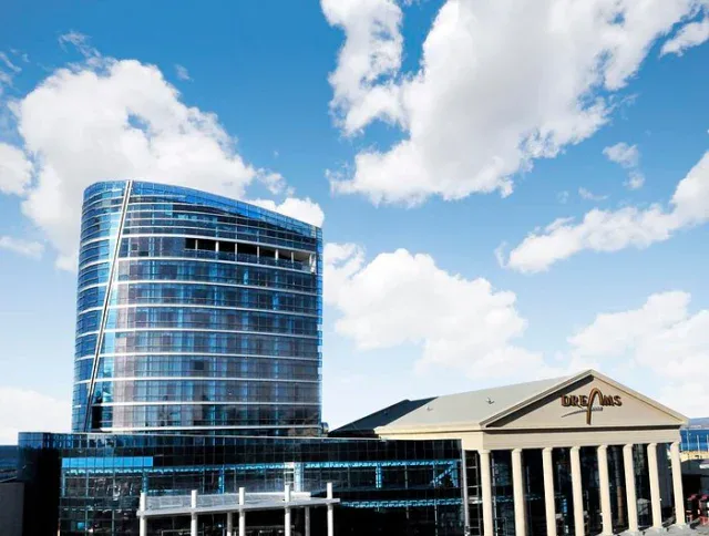A modern glass high-rise building stands beside a classical-style structure with columns under a partly cloudy blue sky.