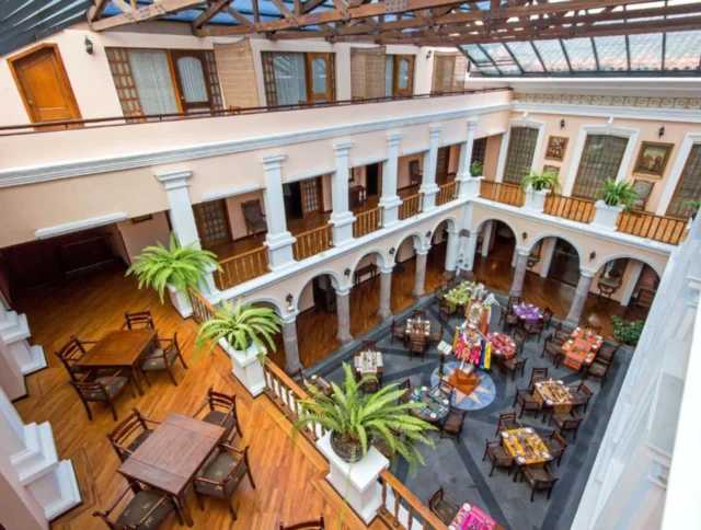 A large, bright atrium with plants, wooden tables, colorful decorations, and a glass ceiling, surrounded by two levels of arched balconies.