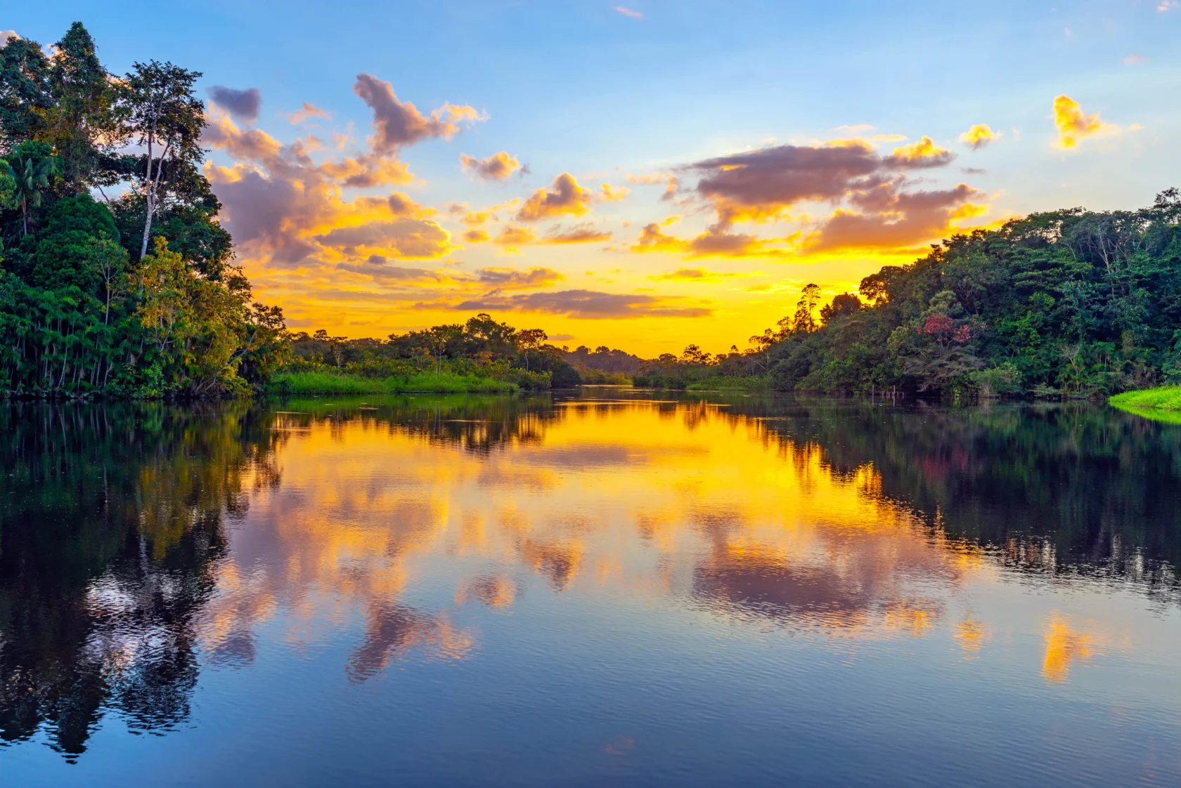 Sunset over a calm river with lush greenery and trees on both sides, reflecting vibrant colors of orange, pink, and blue in the sky and water.