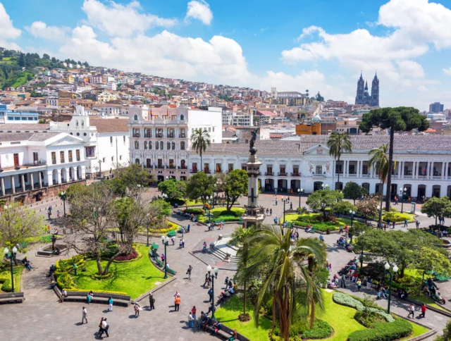 Aerial view of a bustling city square with a central statue, surrounded by manicured gardens, historical buildings, and a hillside backdrop under a blue sky.