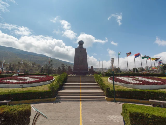 Monument with a globe on top, surrounded by gardens and flags, under a partly cloudy sky.