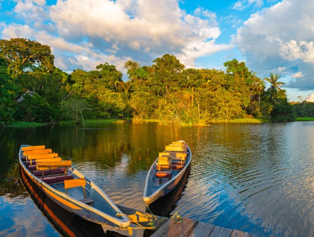 Two wooden boats are docked on a calm river surrounded by lush green forest under a partly cloudy sky.