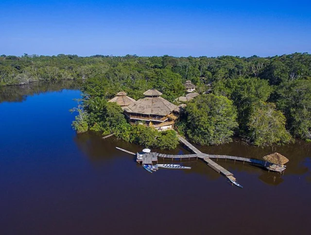 Aerial view of a resort with thatched-roof buildings surrounded by dense forest and a brown river. A wooden walkway leads to small docks with boats.