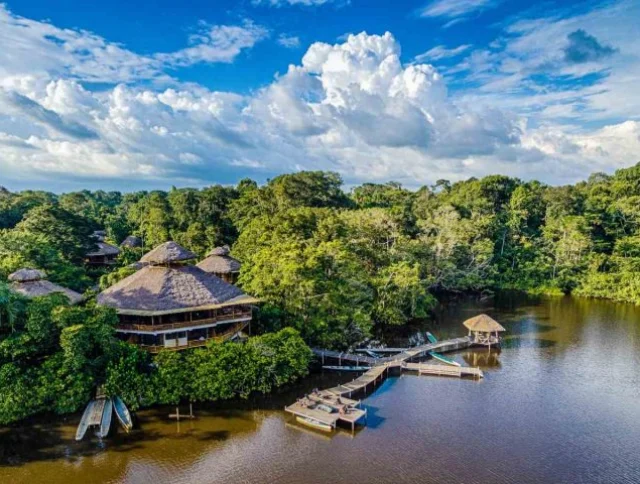 Aerial view of a riverside lodge surrounded by dense trees, featuring thatched buildings, a dock, and a small pavilion on the water, under a partly cloudy sky.