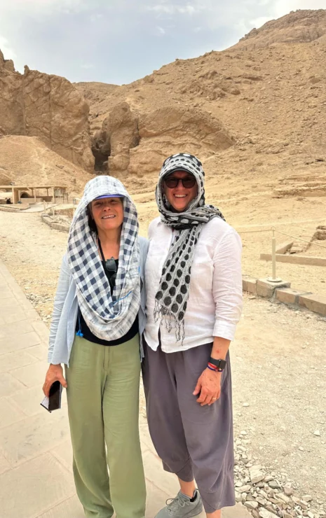 Two people standing on a paved path in a rocky, desert-like area. They are wearing light clothing and scarves around their heads, smiling at the camera.