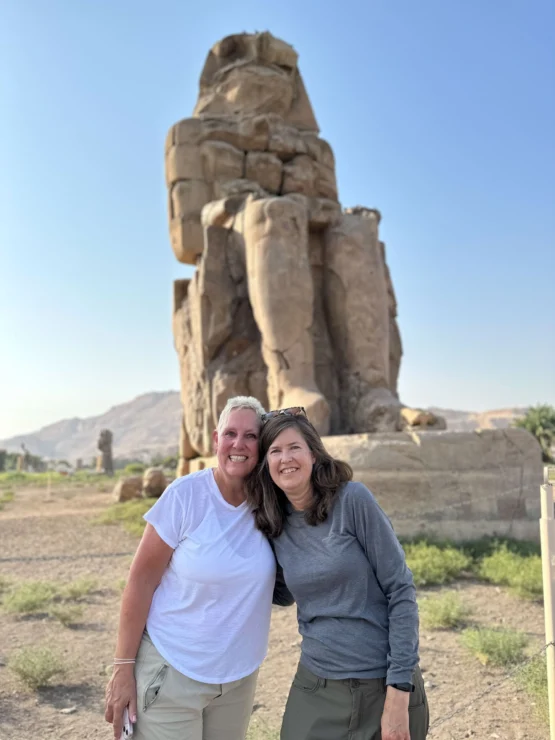 Two people standing in front of a large ancient stone statue in a desert landscape under a clear blue sky.