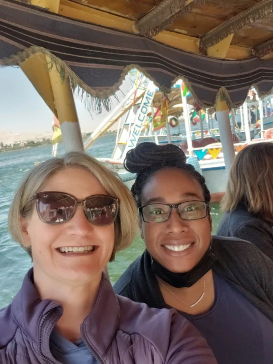 Two people smiling on a boat with colorful decorations on a sunny day. A "Welcome" sign is visible in the background.