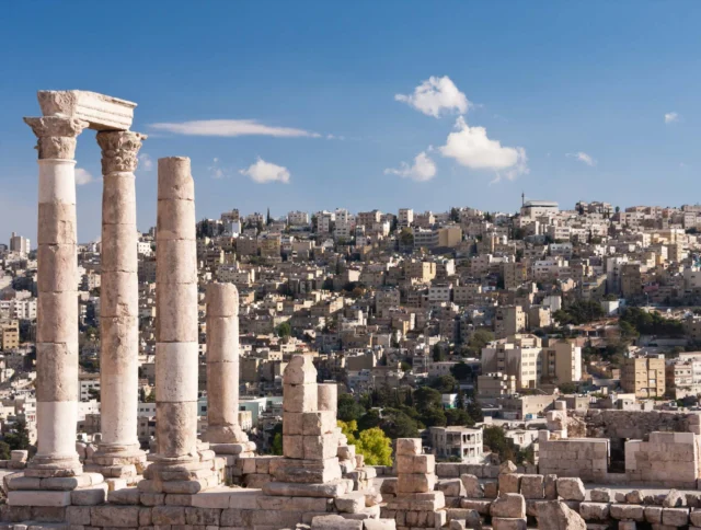 Four ancient columns stand against a backdrop of a modern cityscape under a blue sky with scattered clouds.