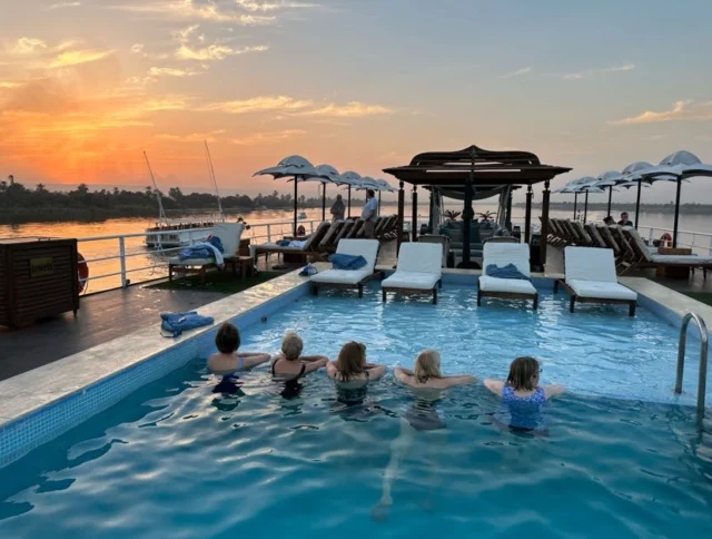 Five people relax in a pool on a boat deck at sunset, overlooking a serene river. Lounge chairs and umbrellas line the deck.