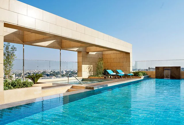 Rooftop swimming pool with clear blue water, two lounge chairs, plants, and a cityscape view in the background.