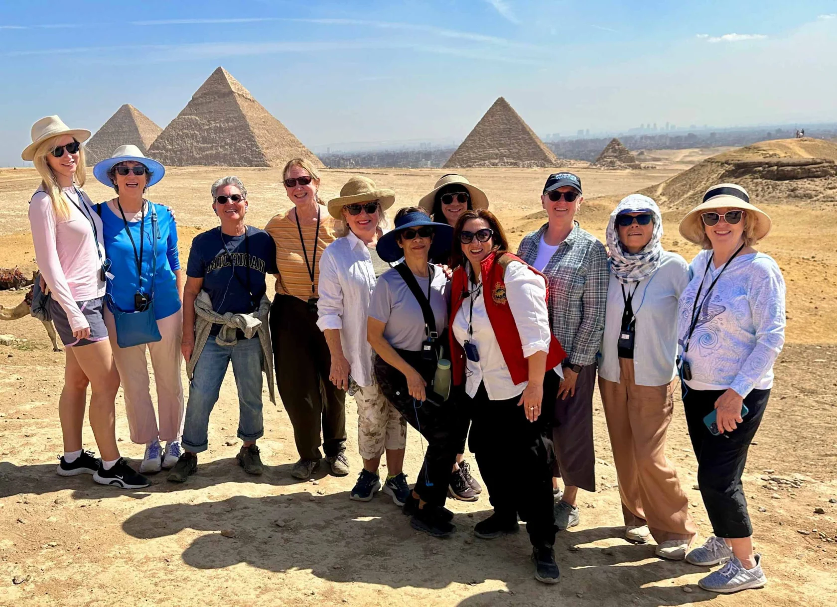 A group of people standing in front of the pyramids in the desert, wearing casual clothing and sun hats, smiling at the camera.