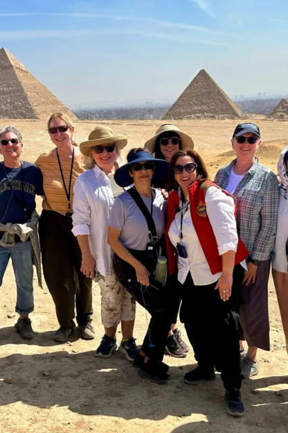 A group of people standing in front of the pyramids in the desert, wearing casual clothing and sun hats, smiling at the camera.