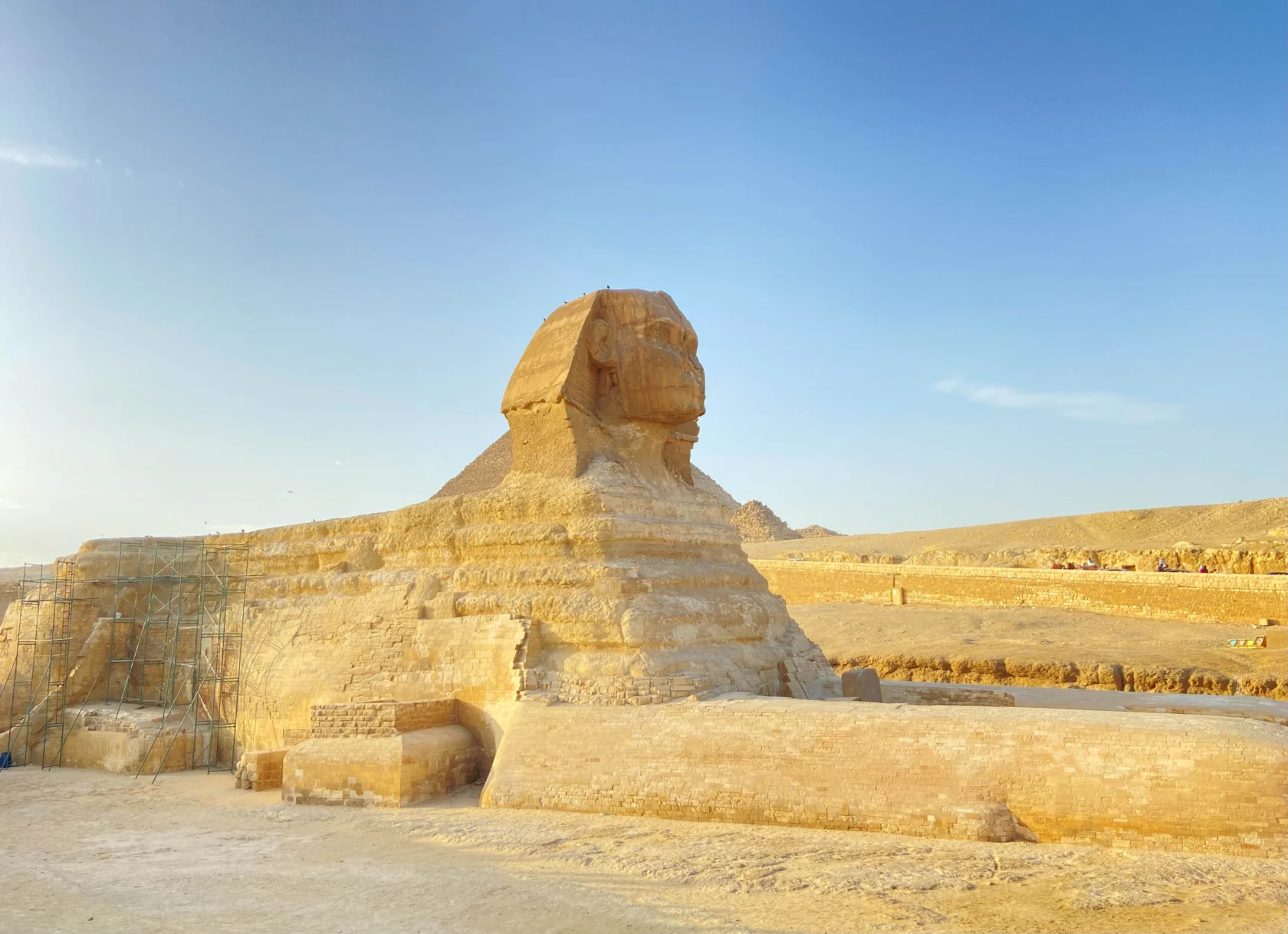 The Sphinx in Egypt, a limestone statue with a lion's body and human head, is seen against a clear blue sky. Scaffolding is present on one side for restoration work.