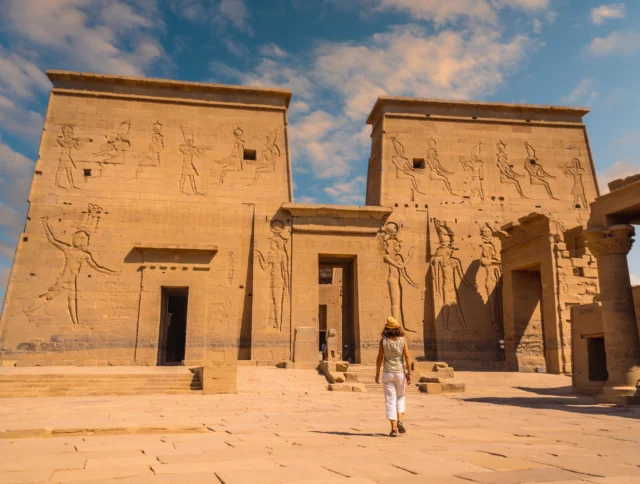 A person walks towards the ancient Philae Temple with tall stone walls adorned with hieroglyphs under a partly cloudy sky.