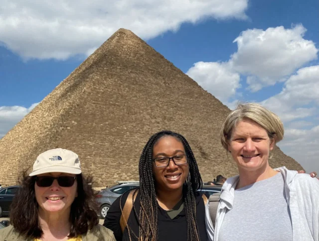 Three people stand in front of a large pyramid under a partly cloudy sky.