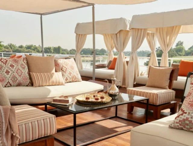 Luxurious outdoor lounge area with cushioned sofas, patterned pillows, a glass coffee table with snacks, and canopies on a boat deck overlooking greenery and water.