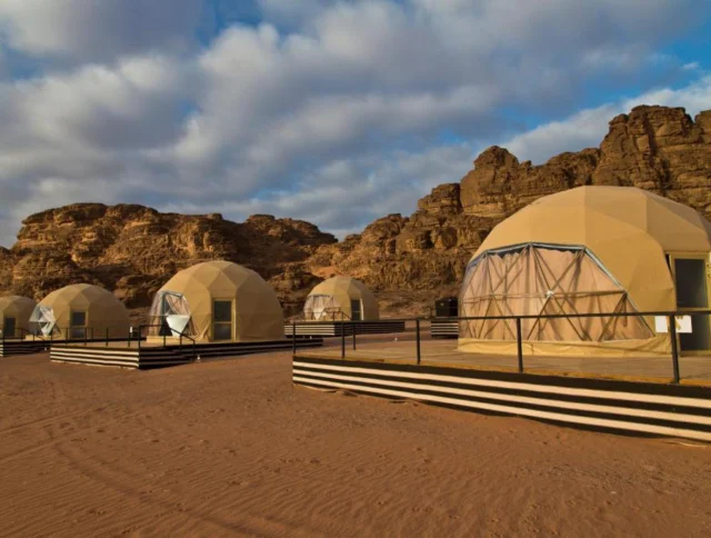 A row of dome-shaped tents on a sandy landscape, with rocky cliffs in the background under a partly cloudy sky.