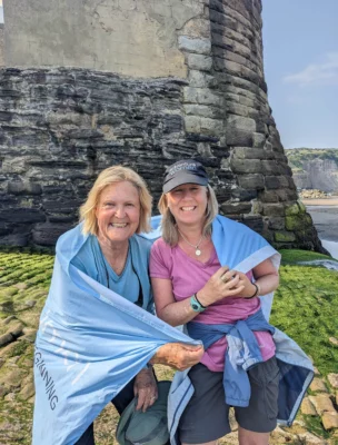 Two women smiling and standing together, wrapped in a blue cloth, in front of a stone tower on a rocky outdoor setting.