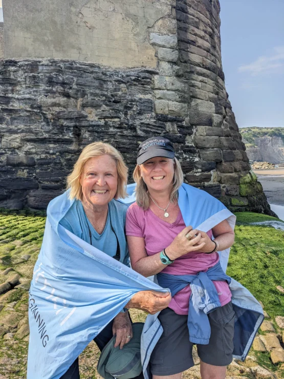 Two women smiling and standing together, wrapped in a blue cloth, in front of a stone tower on a rocky outdoor setting.