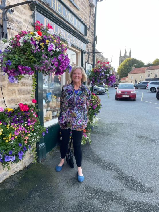 A person stands smiling in front of a delicatessen with hanging baskets of colorful flowers. Cars and a church tower are visible in the background.