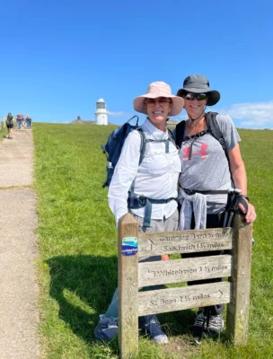 Two people in hiking attire stand behind a wooden signpost on a grassy path, with a lighthouse in the background.