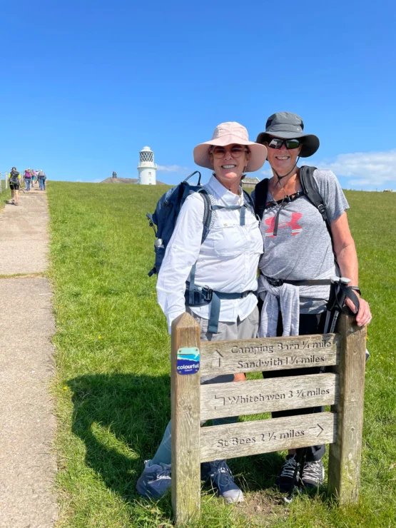 Two people in hiking attire stand behind a wooden signpost on a grassy path, with a lighthouse in the background.