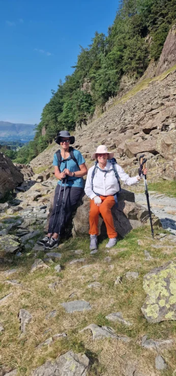 Two people in hiking gear rest on rocks, holding trekking poles. They are surrounded by rocky terrain, with trees and a clear blue sky in the background.