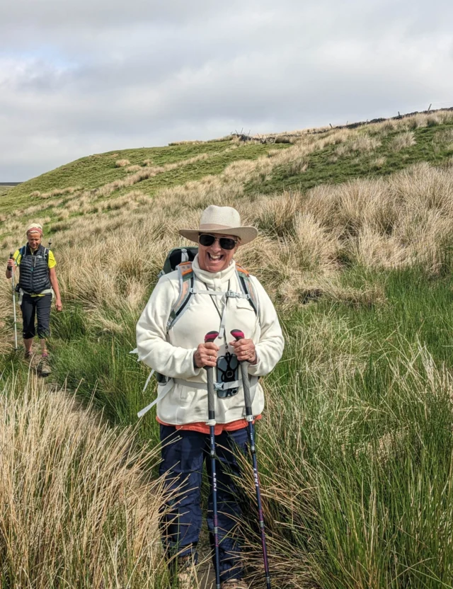 Two people hiking through grassy terrain, one wearing a hat and holding trekking poles, and another following behind.