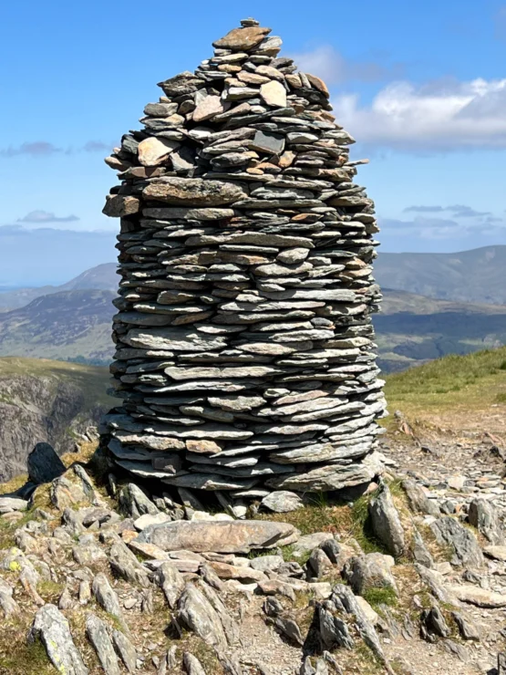 A tall cairn made of stacked stones is situated on a rocky hilltop under a clear blue sky, with distant mountains in the background.