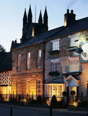 Exterior of a warmly lit hotel at dusk, with a mix of stone and timber architecture and towers in the background.