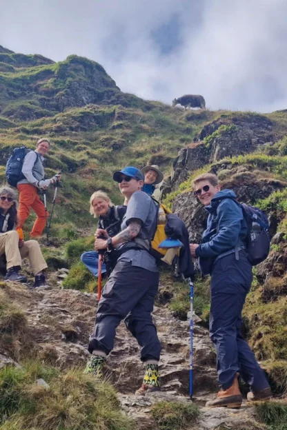 A group of hikers pauses on a grassy, rocky hillside under a partly cloudy sky.