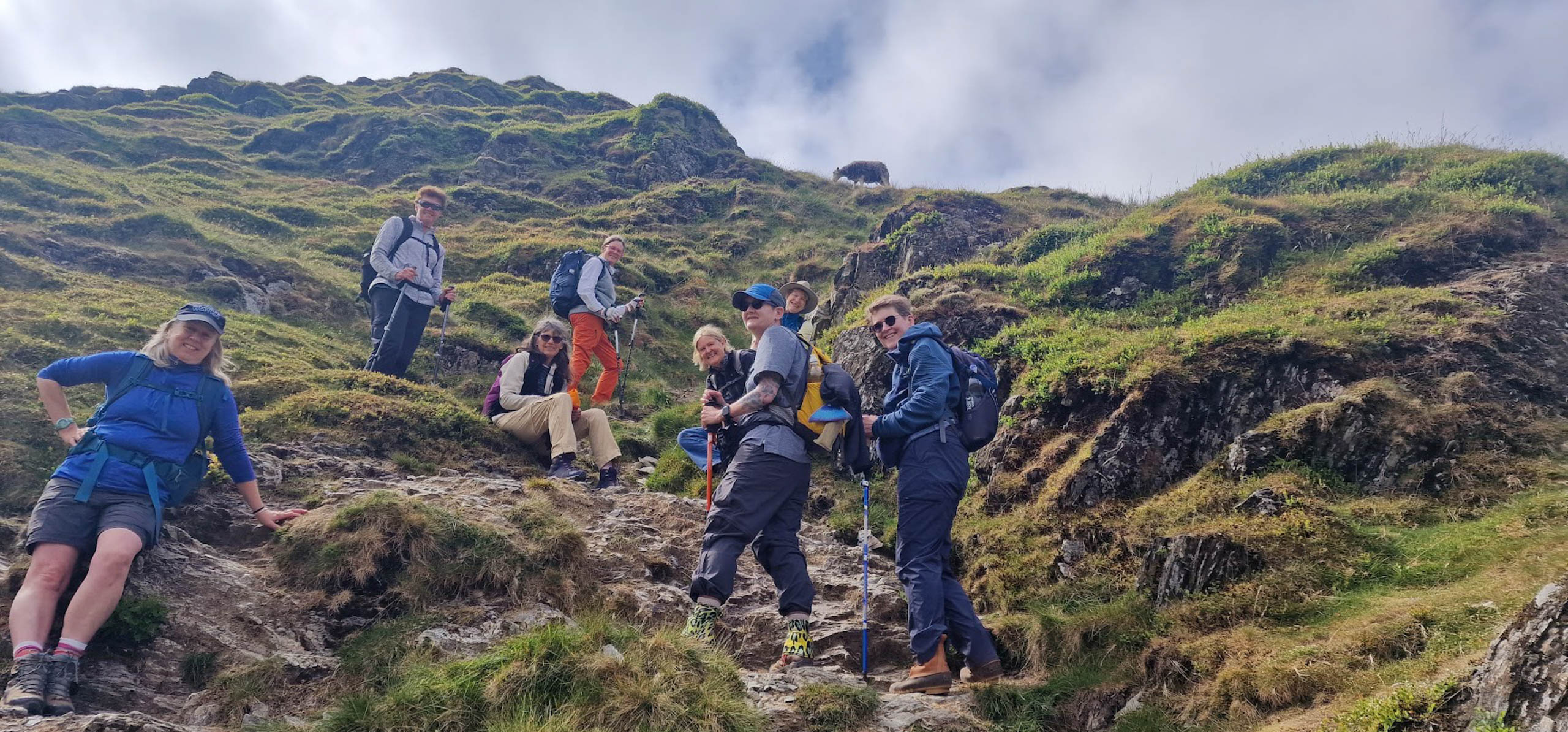 A group of people hiking up a grassy and rocky hillside, dressed in outdoor gear and carrying backpacks.