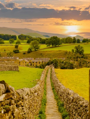 A stone path leads through a lush green landscape toward a village, with a sunset visible on the horizon. Stone walls and trees line the path.