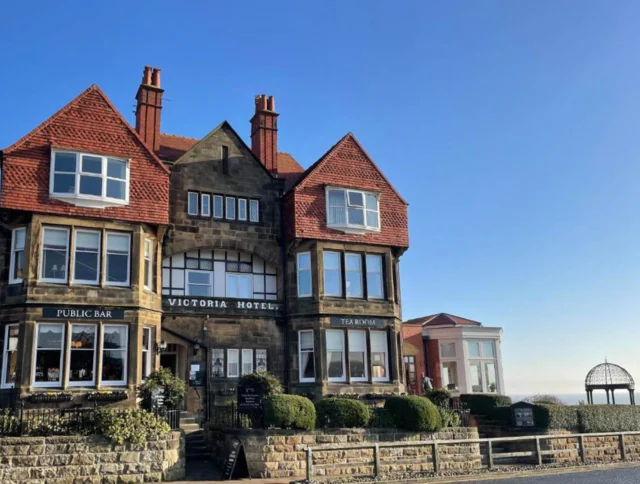 A large stone building with red roofs labeled "Victoria Hotel" featuring a public bar and tearooms, set against a clear blue sky.