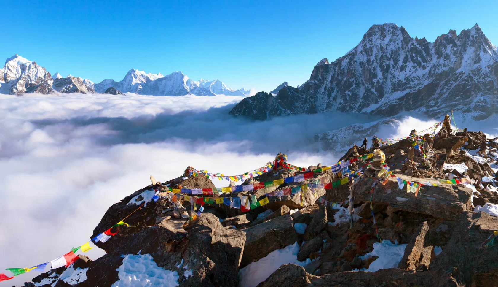 Snow-covered mountains with colorful prayer flags atop rocky terrain, under a clear blue sky, with clouds below.