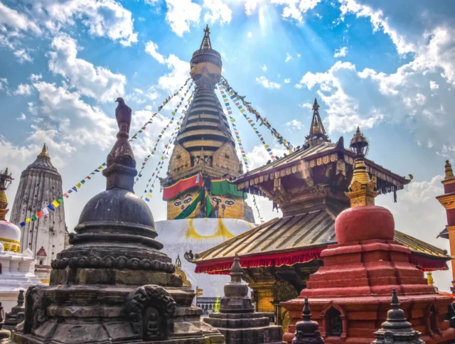 Colorful Buddhist stupa with prayer flags in a temple complex under a bright blue sky with clouds.